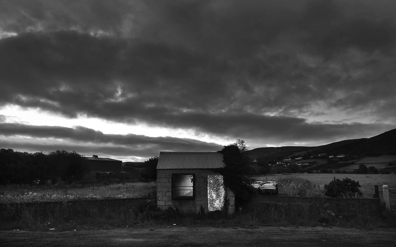 A small abandoned single-room border shack stands beside a road and field with bales of hay and mountains visible in the background.