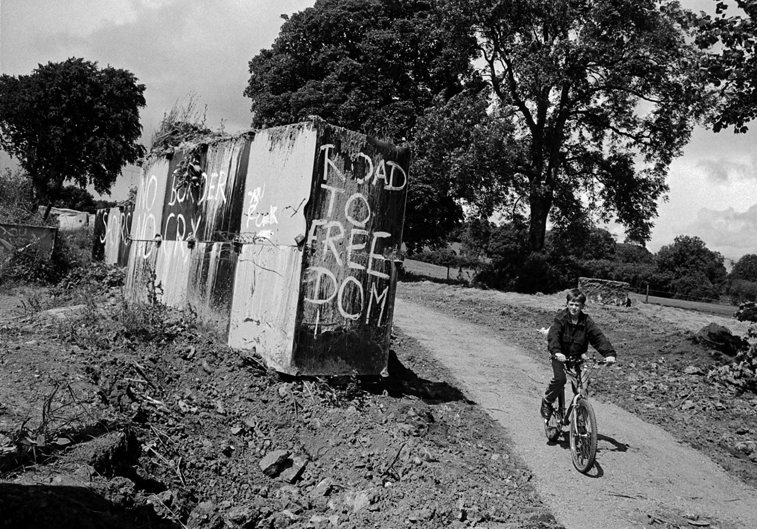 A young boy on his bike cycles on a dirt track past large concrete blocks painted with graffiti criticising the border