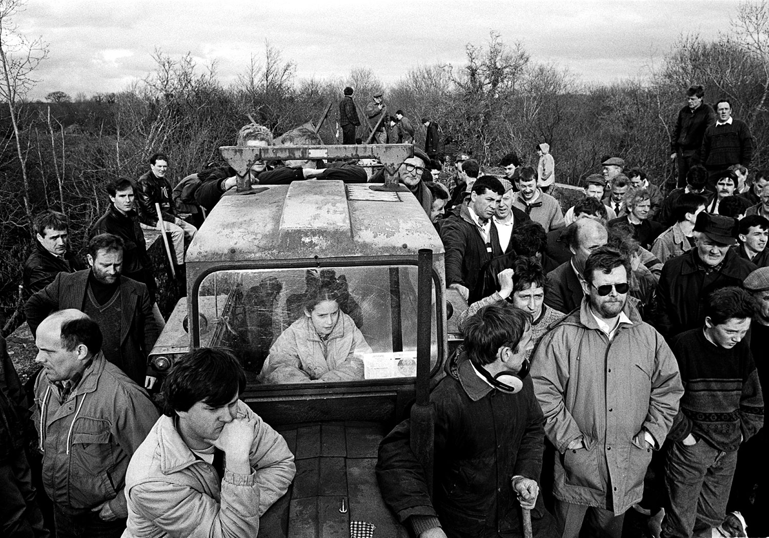 A crowd of men of all ages stand on a barricaded bridge looking at something off-screen. There is a lone girl sitting in a tractor in the middle of the crowd.