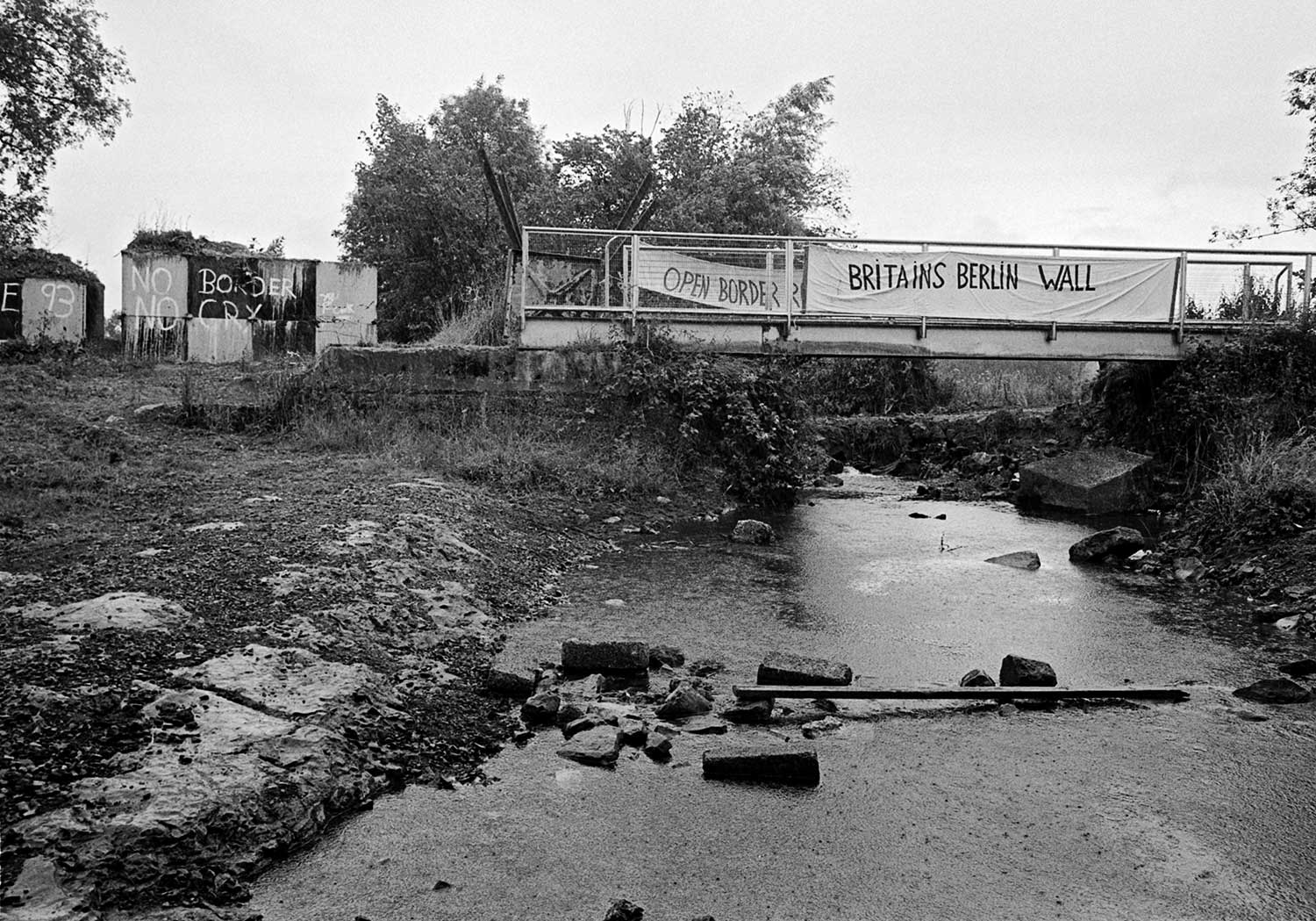 A small makeshift rusty bridge crosses a stream. Banners saying Britain’s Berlin Wall and open border hang from the bridge.