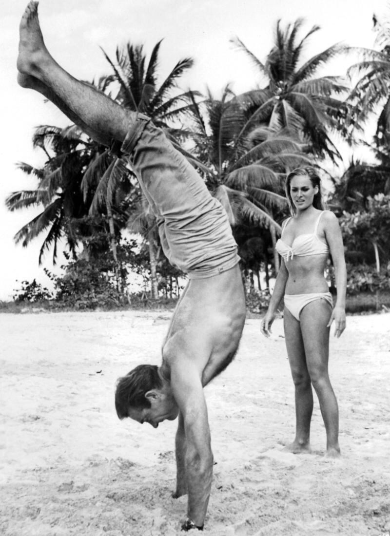 Sean Connery doing a hand stand in the sand while Ursula Andress watches in amazement in a scene from the film James Bond: Dr No, 1962. Photograph: United Artist/Getty