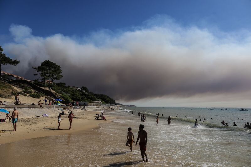 Beach-goers bathe and lay at a beach of "Pyla sur mer" as a black cloud of smoke from a fire that hit La Teste-de-Buch forest rises from the Pyla Dune in the background, in the Arcachon basin southwest France, on July 13, 2022. - French Interior Minister said "2.800 hectares of pine trees have gone to smoke", as two fires hit the Gironde department, amid a heatwave across the country. Photo by Thibaud Moritz / AFP via Getty Images)