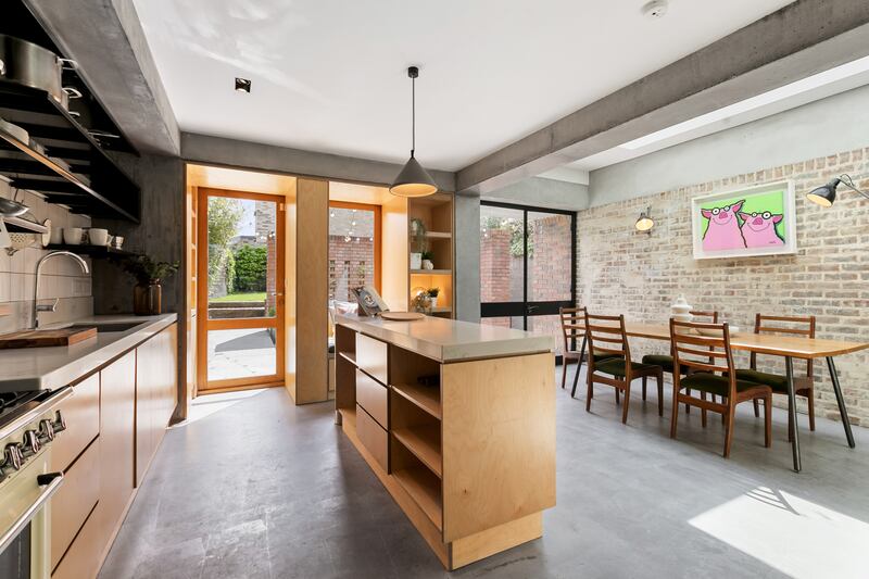 A contemporary kitchen of beech ply has concrete flooring and an aged red brick wall for interest